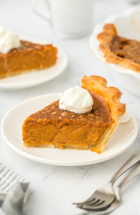 A slice of pumpkin pie on a plate topped with whipped cream with a fork next to it and more slices on the background