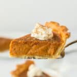 A pie server holding a slice of pumpkin pie with whipped cream on top