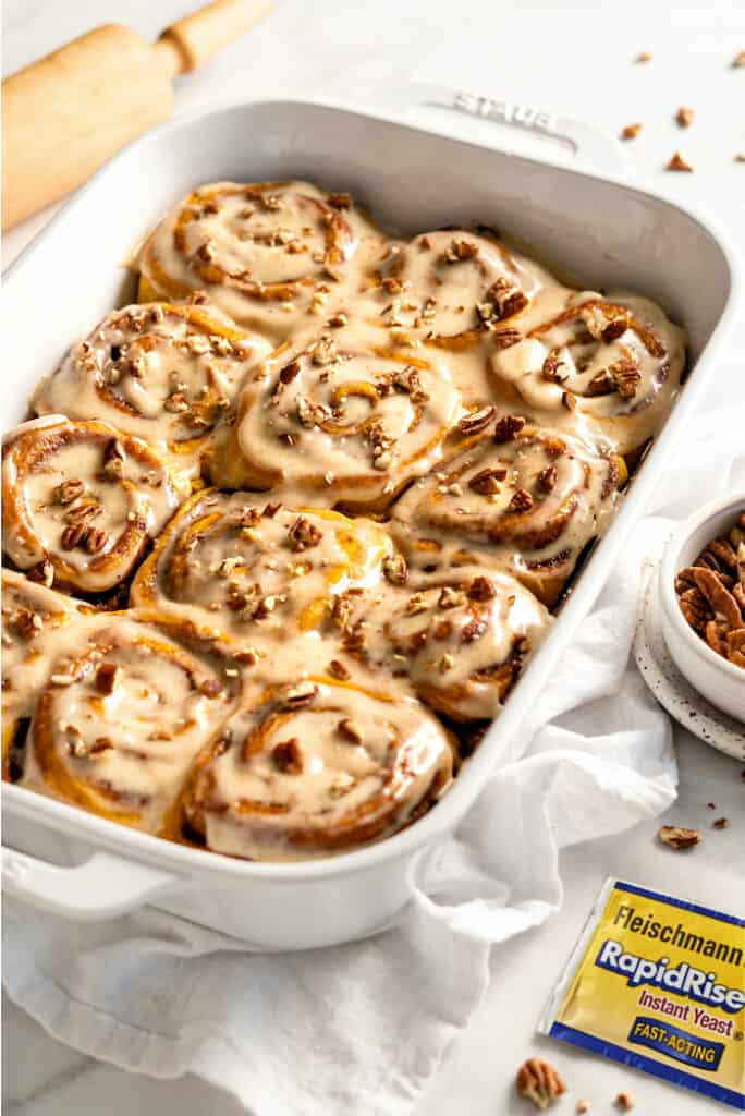 A white casserole dish filled with sweet potato cinnamon rolls against white background with pecans in a white bowl