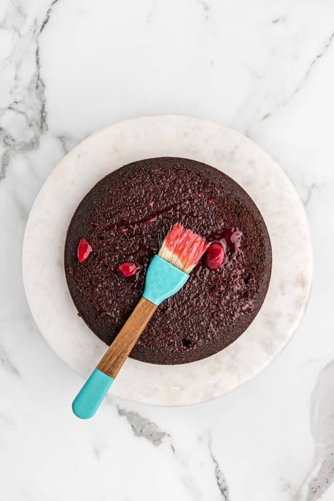 First layer of chocolate cake on the counter with a pastry brush on top brushing cherry sauce