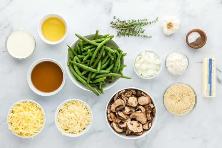 Green beans, mushrooms, onions, thyme, spices and cheeses in white bowls on white countertop