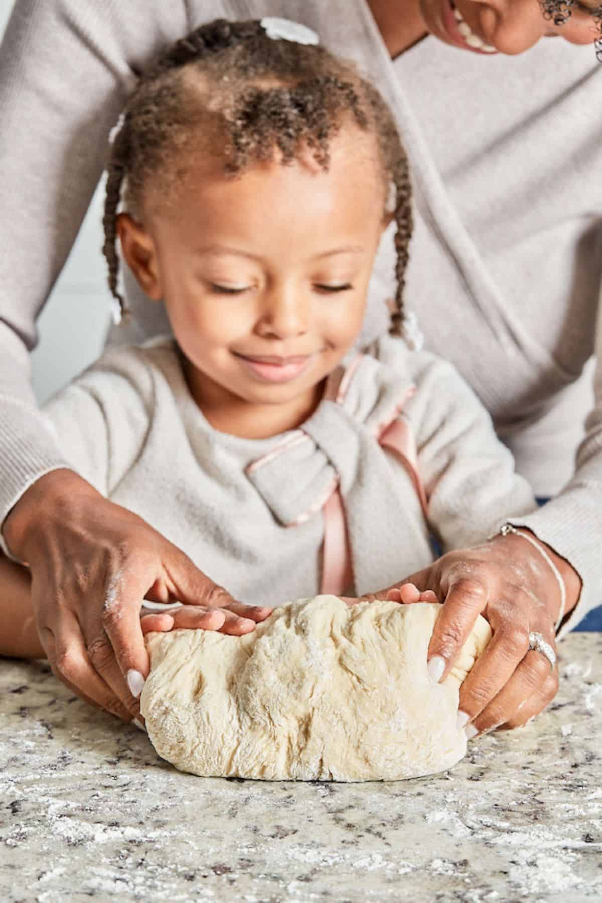 A close-up of a young child smiling while kneading dough with the guidance of an adult’s hands