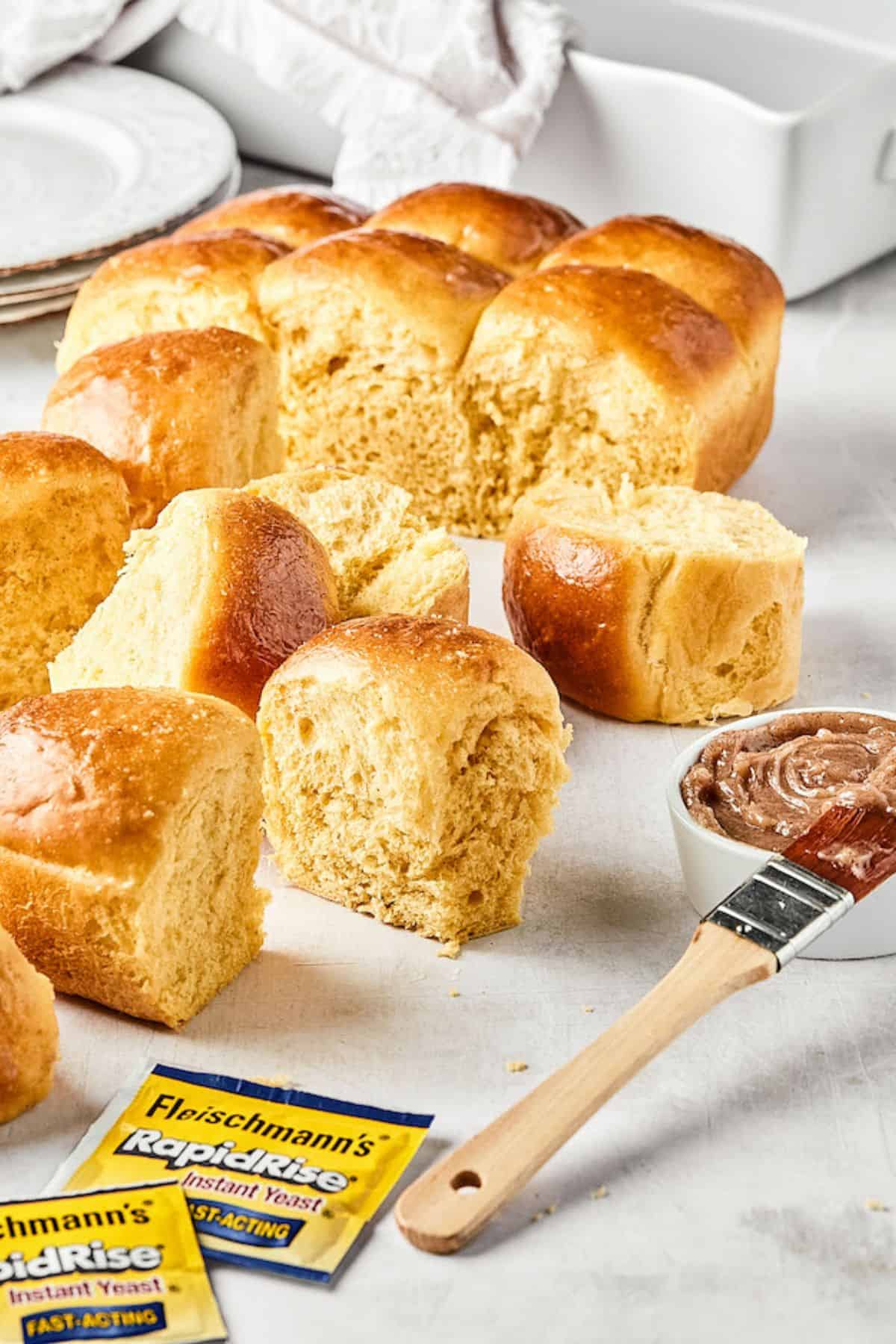 A baking dish of honey butter dinner rolls with packets of yeast and a brush dipped in cinnamon butter on the side