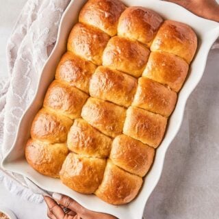 A baking dish with evenly arranged, freshly baked honey butter dinner rolls, being held by two hands.