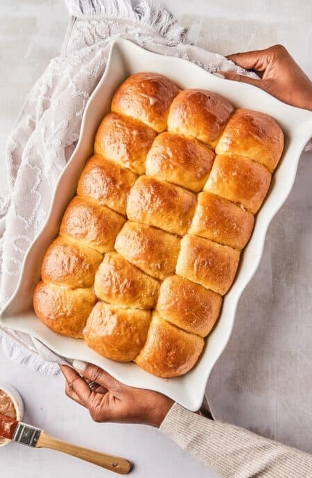 A baking dish with evenly arranged, freshly baked honey butter dinner rolls, being held by two hands.