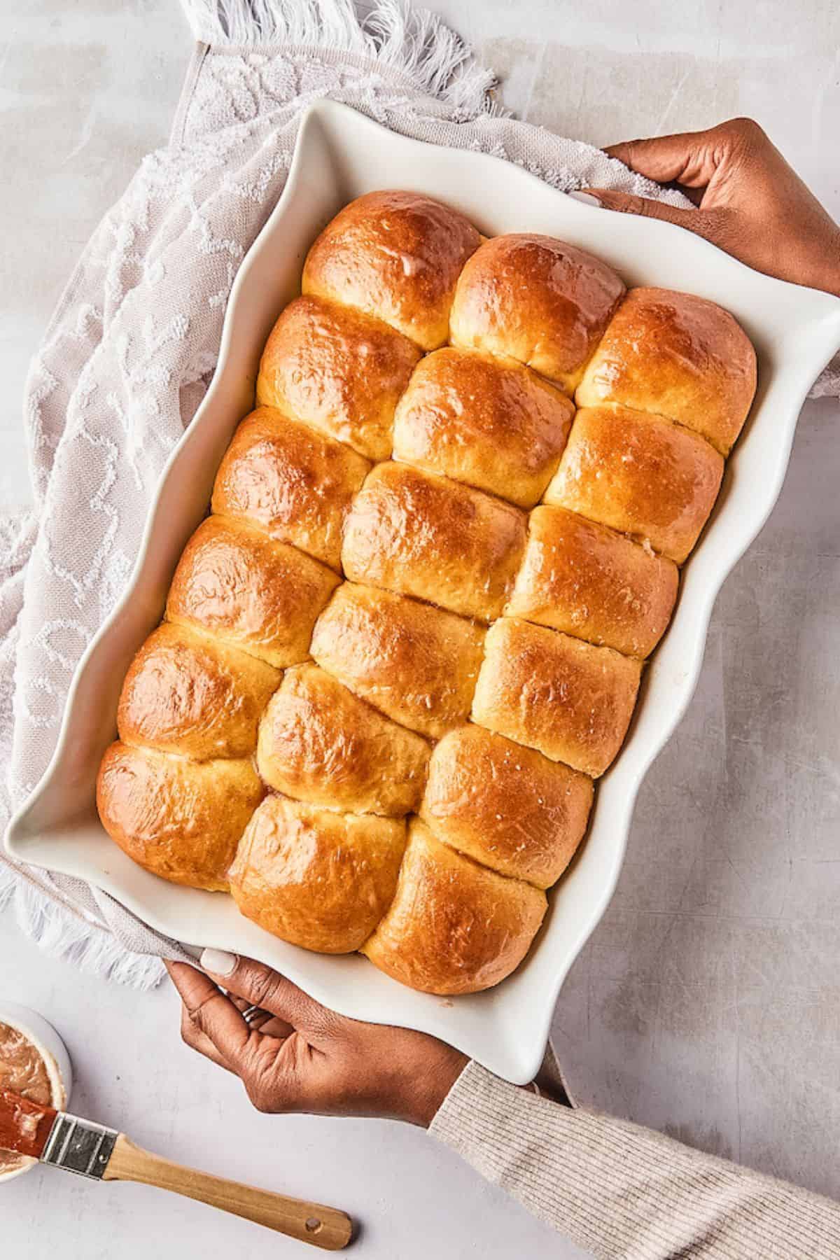 A baking dish with evenly arranged, freshly baked honey butter dinner rolls, being held by two hands.