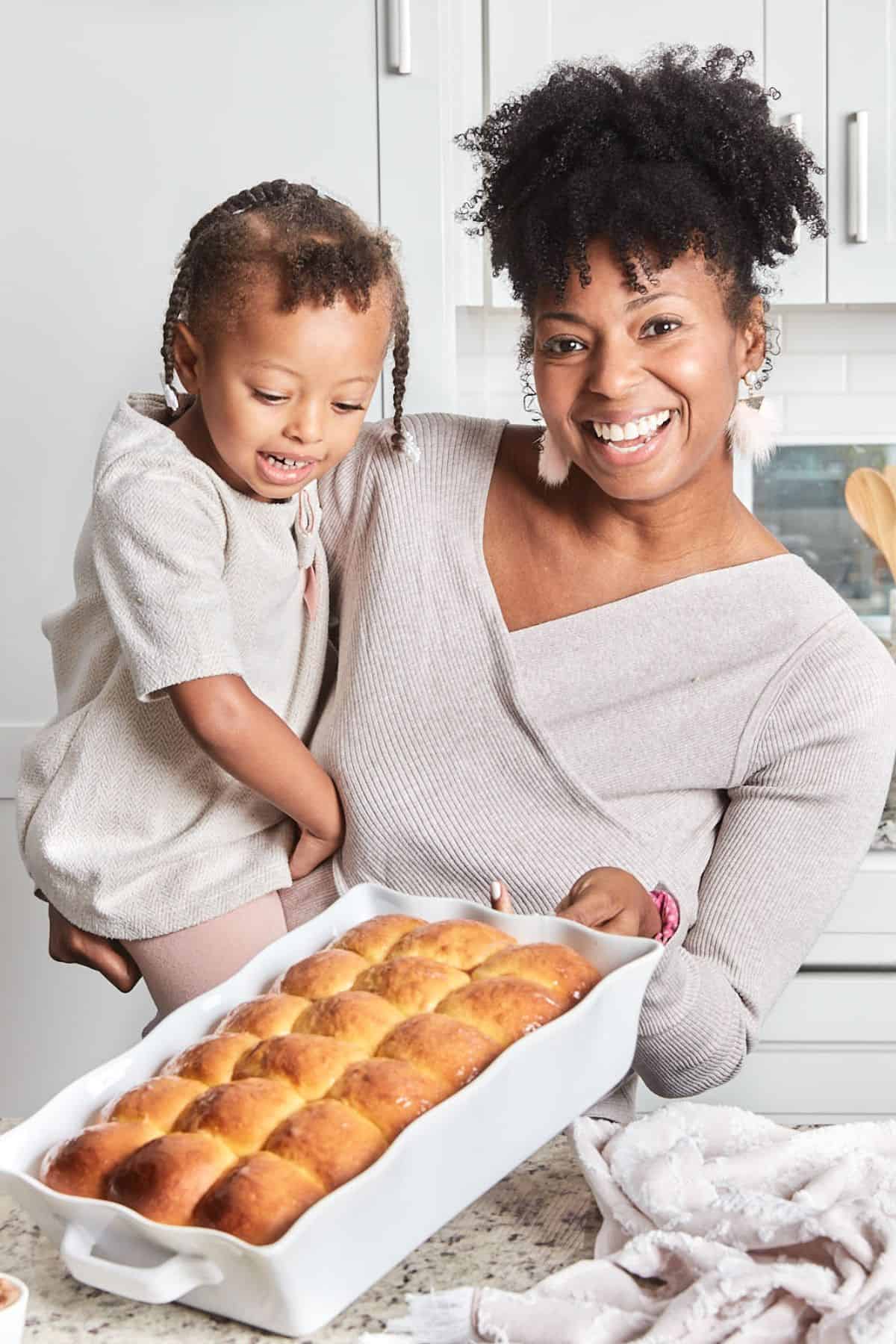 A woman and a child holding a baking dish of honey butter dinner rolls, both smiling in a kitchen setting