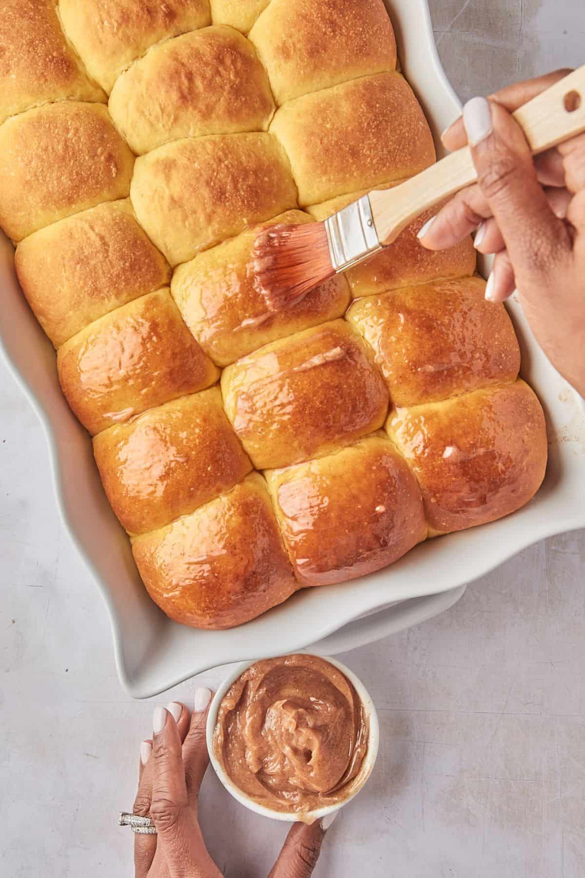 Close-up of a hand brushing melted butter on the tops of warm dinner rolls, with a bowl of cinnamon butter nearby