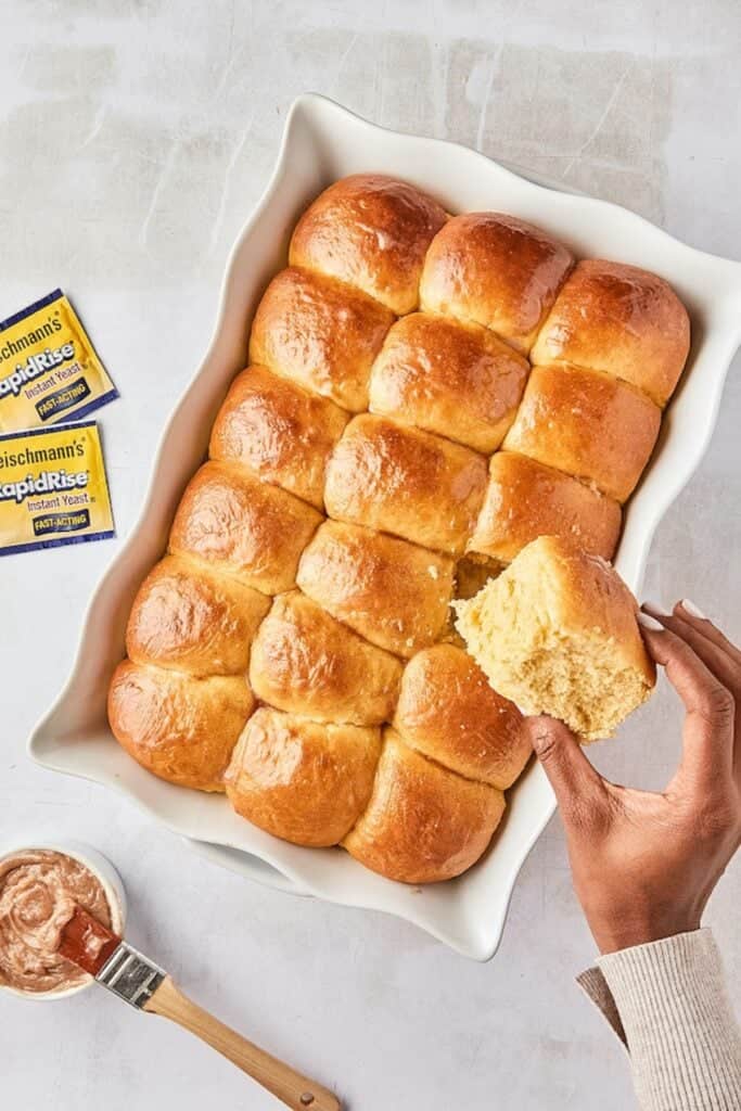 Close-up of a hand breaking off a single dinner roll from a dish filled with baked rolls, ready to eat