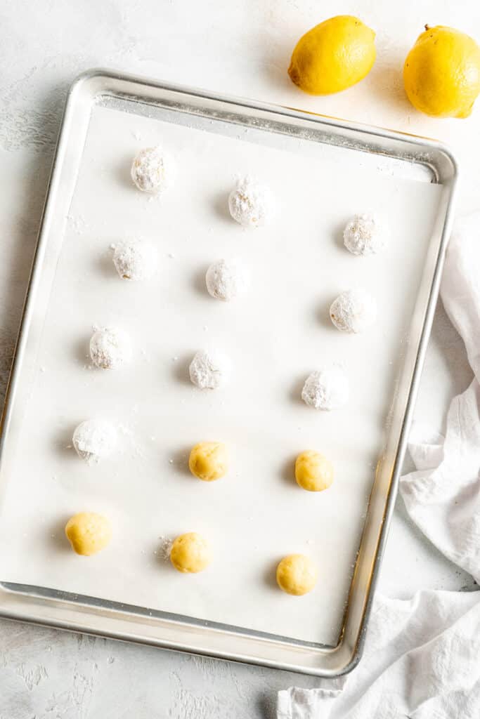 Balls of cookie dough with some that have been rolled in confectioner's sugar on a baking sheet
