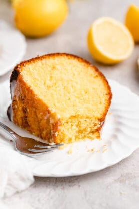A slice of lemon cake on a white plate with a fork cutting into it and lemons around it