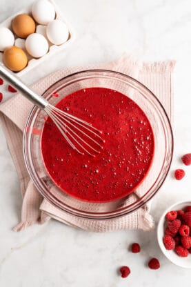 Red velvet cake batter in a clear bowl with a silver whisk