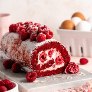 A beautiful raspberry stuffed Red Velvet Cake Roll with raspberries on top against a light pink background