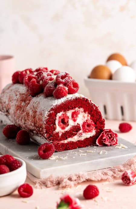 A beautiful raspberry stuffed Red Velvet Cake Roll with raspberries on top against a light pink background