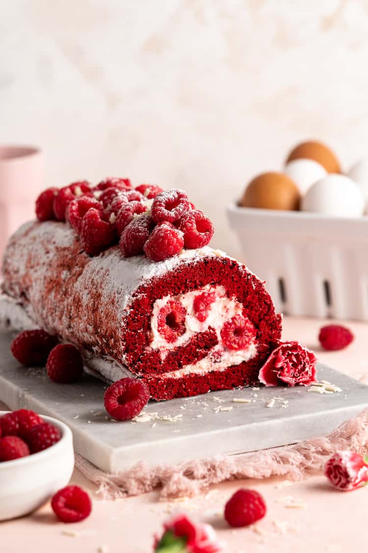 A beautiful raspberry stuffed Red Velvet Cake Roll with raspberries on top against a light pink background