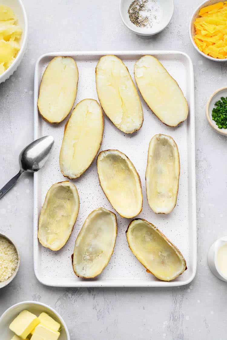 Sliced baked potatoes with filling scooped out on baking sheet