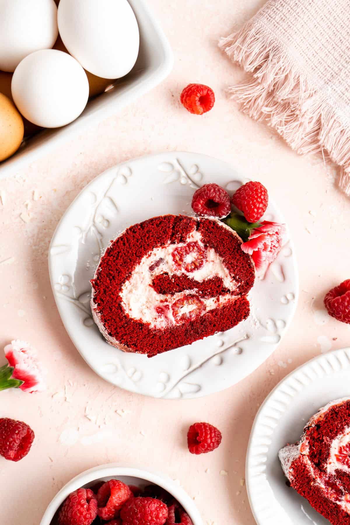 A slice of red velvet cake roll on a white plate with raspberries and eggs next to it