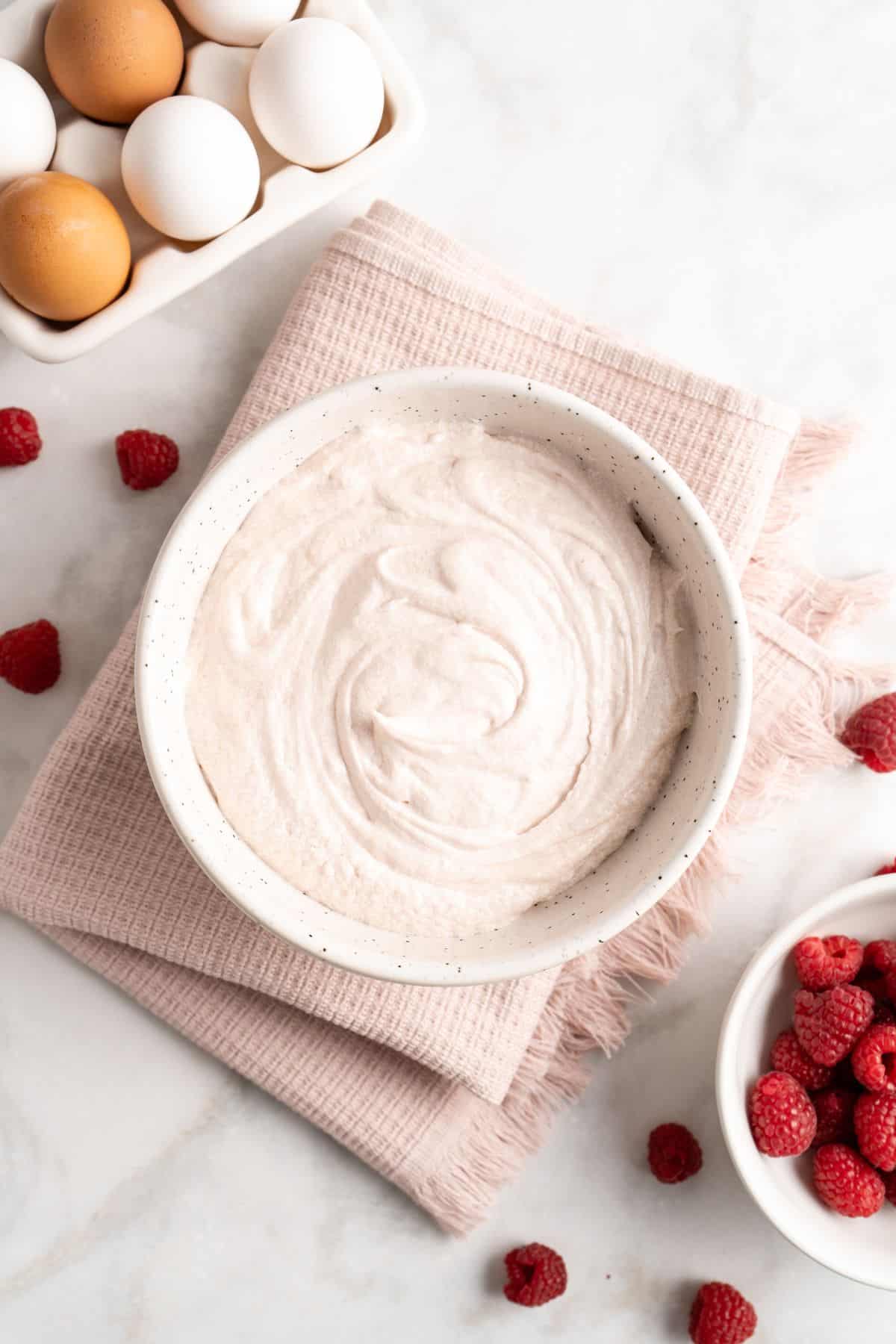 A bowl of creamy buttercream with a pale pink tint, surrounded by fresh raspberries and a tray of eggs