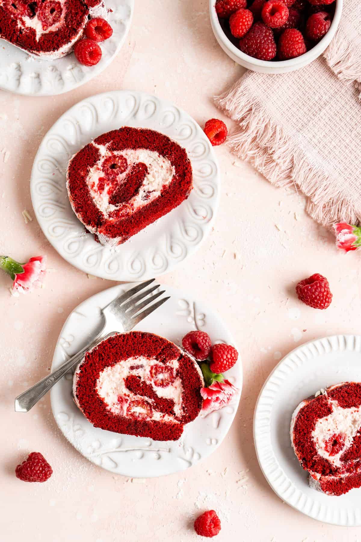 Slices of red velvet cake roll with swirls of pink frosting on plates, garnished with fresh raspberries