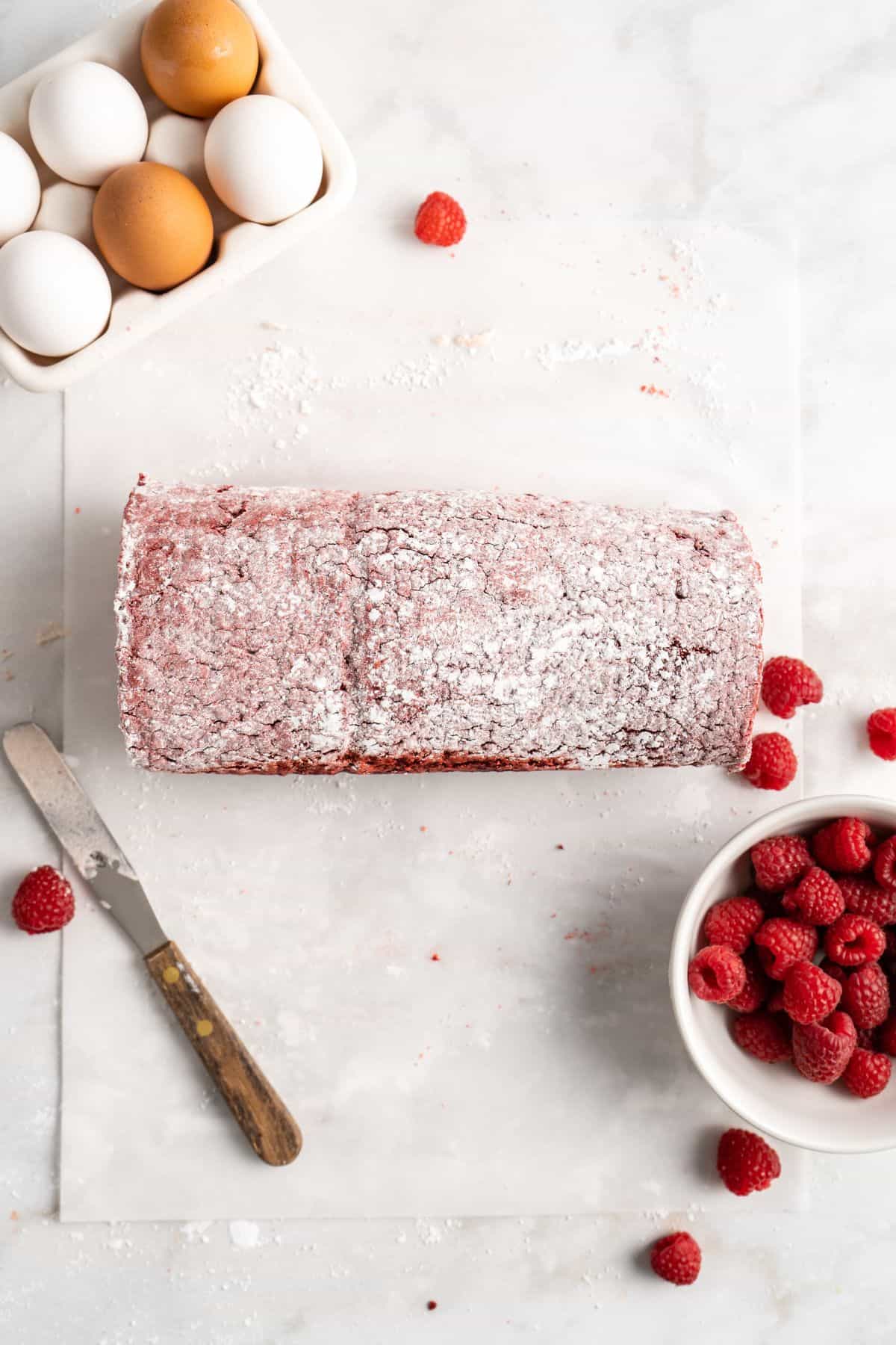 A rolled red velvet cake dusted with powdered sugar, placed on parchment paper with scattered raspberries and a small bowl of raspberries nearby