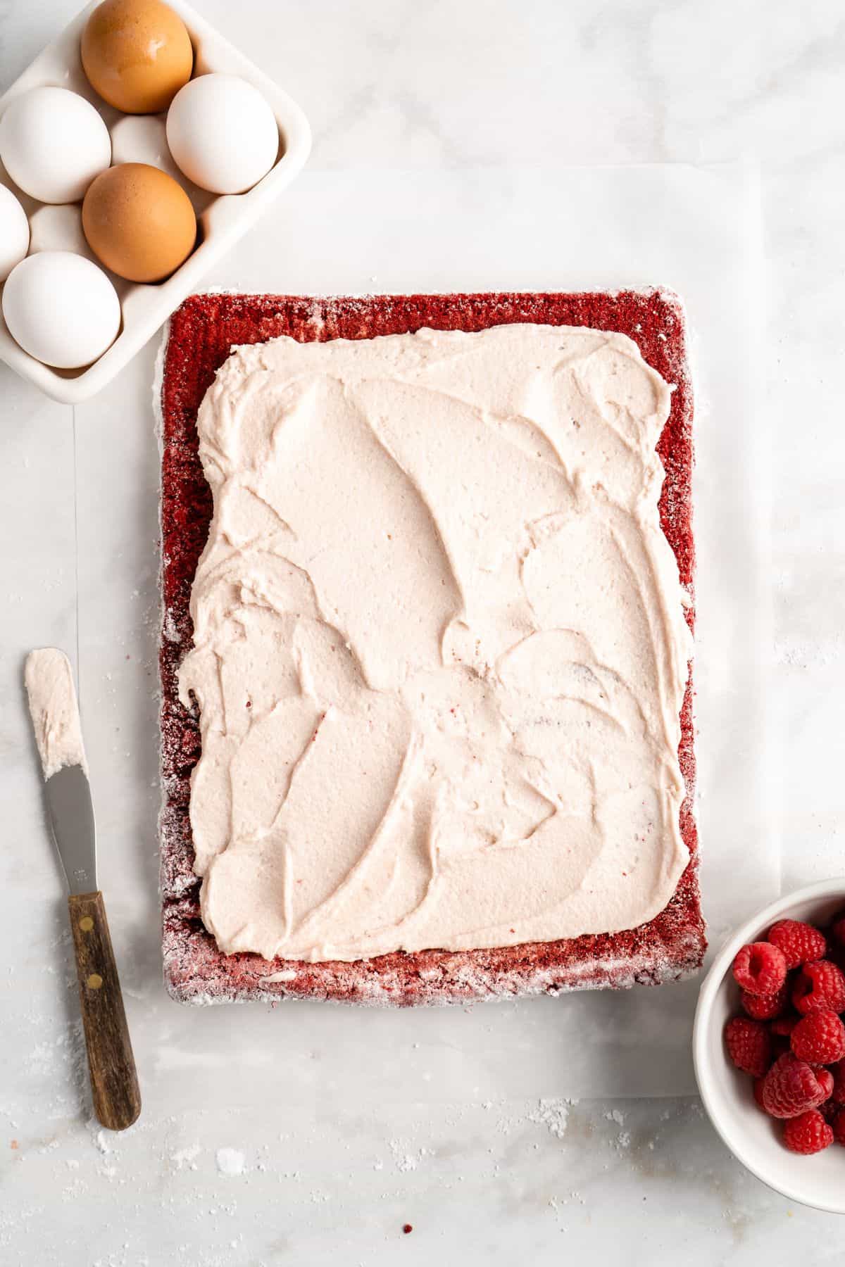 A sheet of red velvet cake partially covered with buttercream, next to a bowl of eggs and fresh raspberries