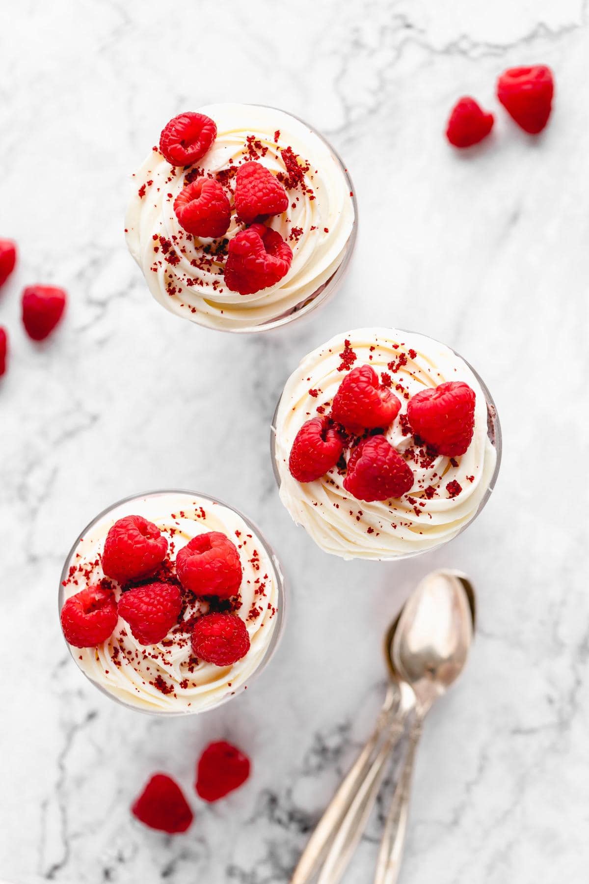 Overhead shot of three red velvet trifles topped with whipped cream and fresh raspberries, with red cake crumbs sprinkled on top