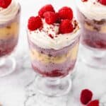 Three red velvet trifles in glass cups showcasing layers of raspberry ice cream, ginger cookies, eggnog sauce, and red velvet cake, topped with whipped cream and raspberries
