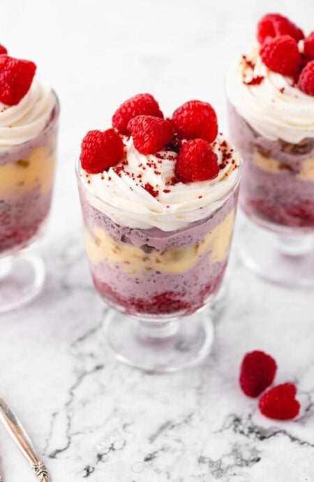 Three red velvet trifles in glass cups showcasing layers of raspberry ice cream, ginger cookies, eggnog sauce, and red velvet cake, topped with whipped cream and raspberries