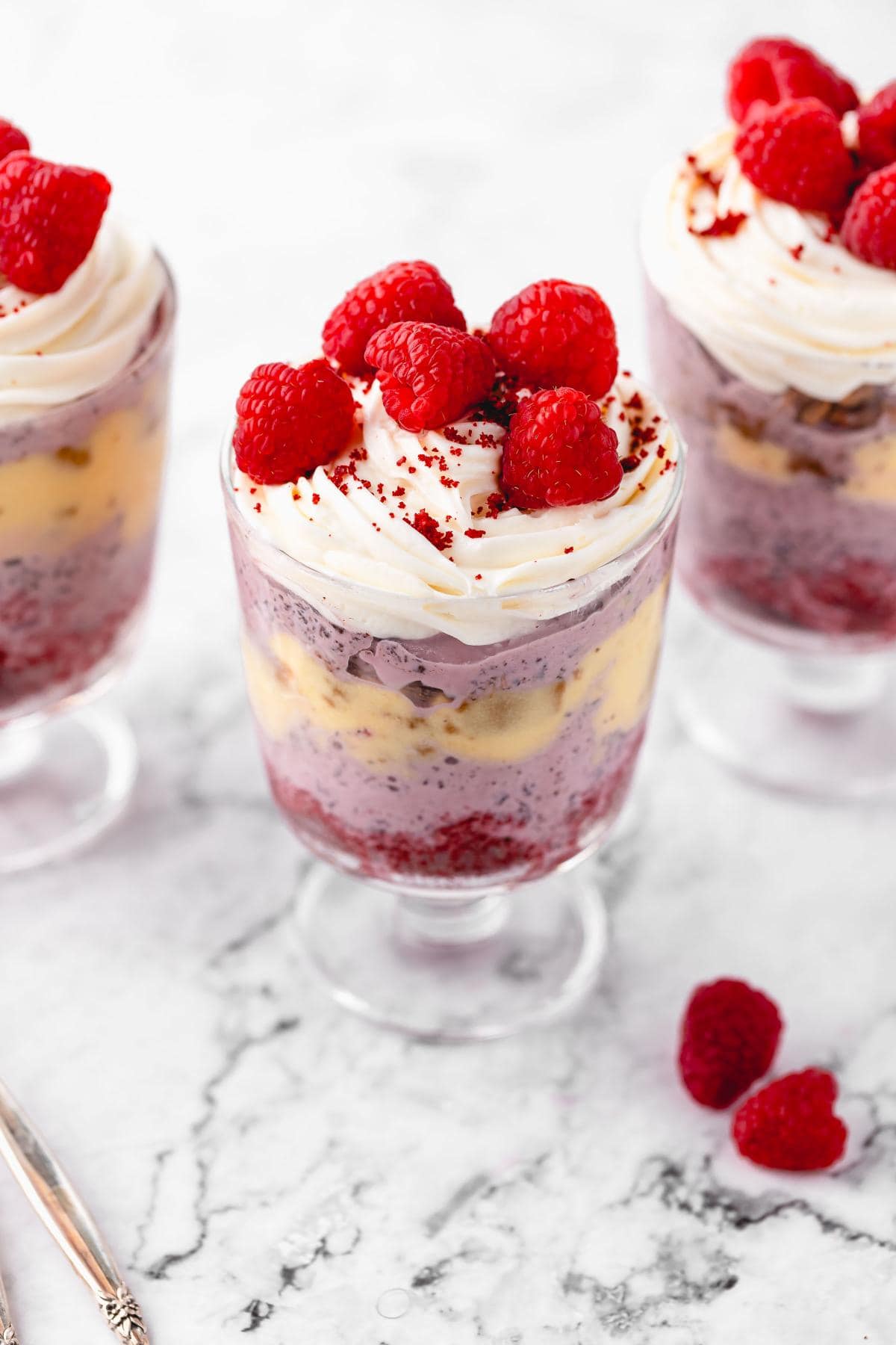 Three red velvet trifles in glass cups showcasing layers of raspberry ice cream, ginger cookies, eggnog sauce, and red velvet cake, topped with whipped cream and raspberries