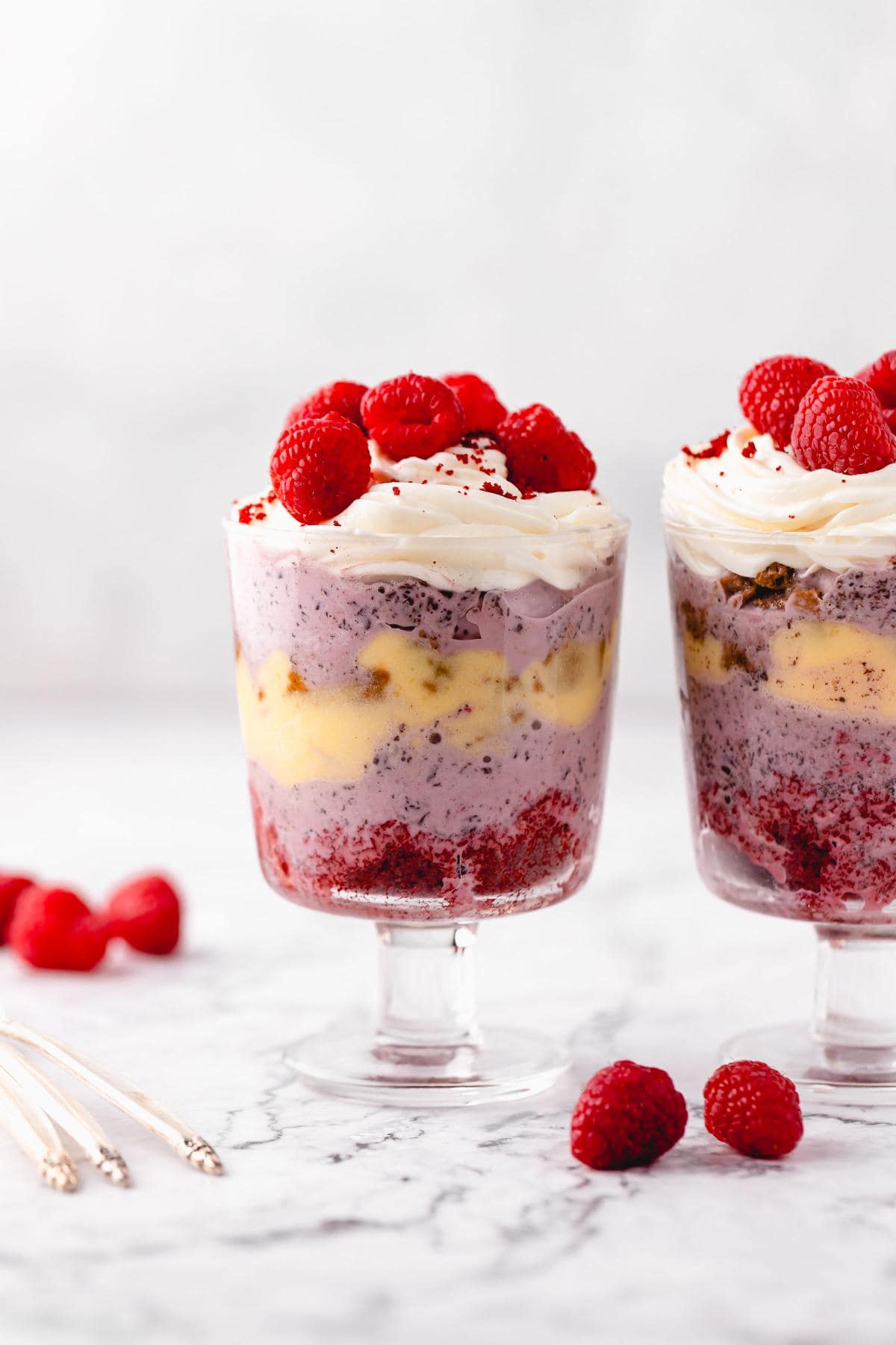 A side-facing shot of two red velvet trifles showing layers of red velvet crumbs, ginger cookies, eggnog sauce and raspberry ice cream, topped with whipped cream and raspberries
