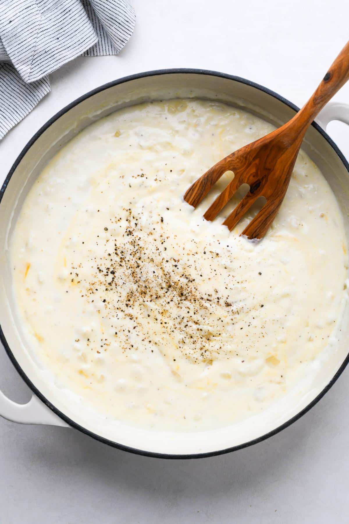 A creamy white sauce with visible black pepper in a pot, stirred with a wooden spatula