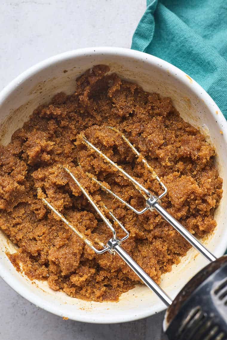 Butter, sugar and molasses being mixed together with a hand mixer in a white bowl with turquoise napkin in background