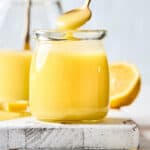 A close up of a jar of homemade lemon curd with a spoon scooping some out