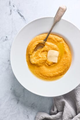 A white bowl filled with sweet potato grits with a pat of butter on top and a spoon mixing everything