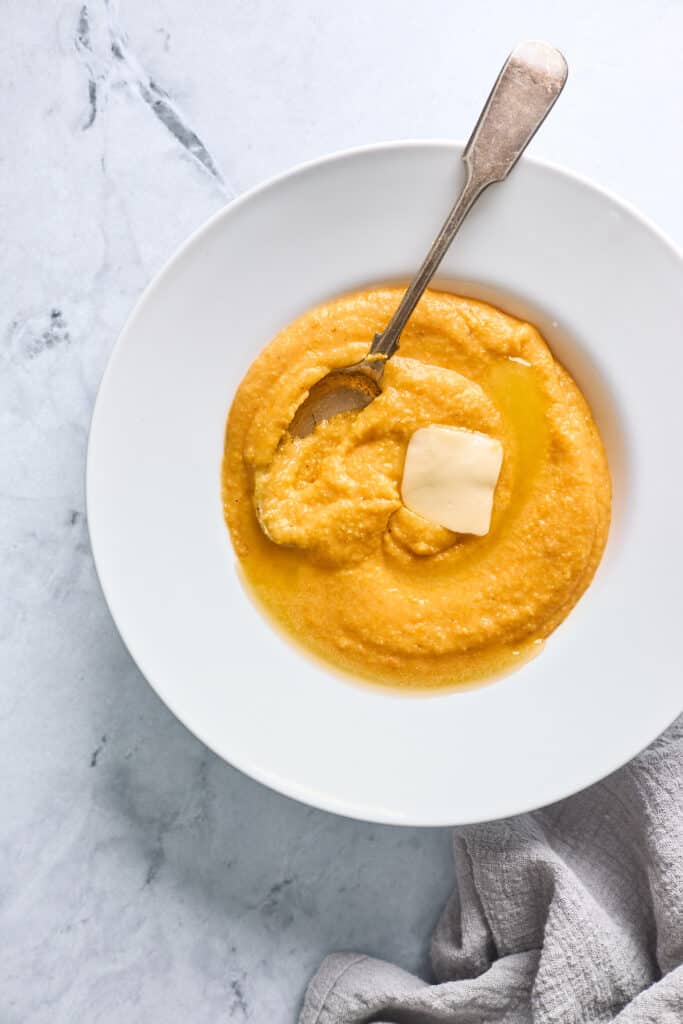 A white bowl filled with sweet potato grits with a pat of butter on top and a spoon mixing everything