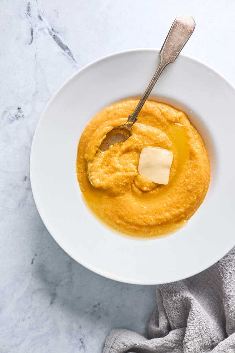 A white bowl filled with sweet potato grits with a pat of butter on top and a spoon mixing everything