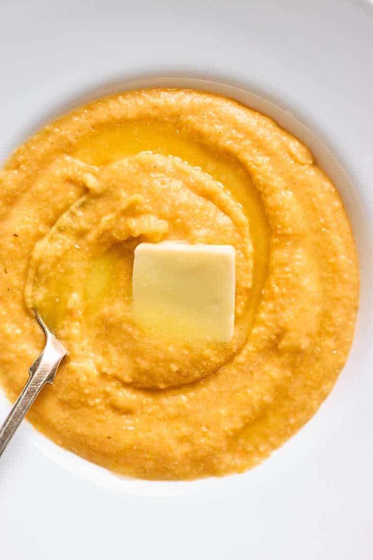 A close up of grits made with sweet potatoes in a white bowl with butter melting over the top