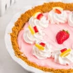A close up of a pink colored kool aid pie recipe topped with whipped cream on a white background with fresh strawberries in the pasta