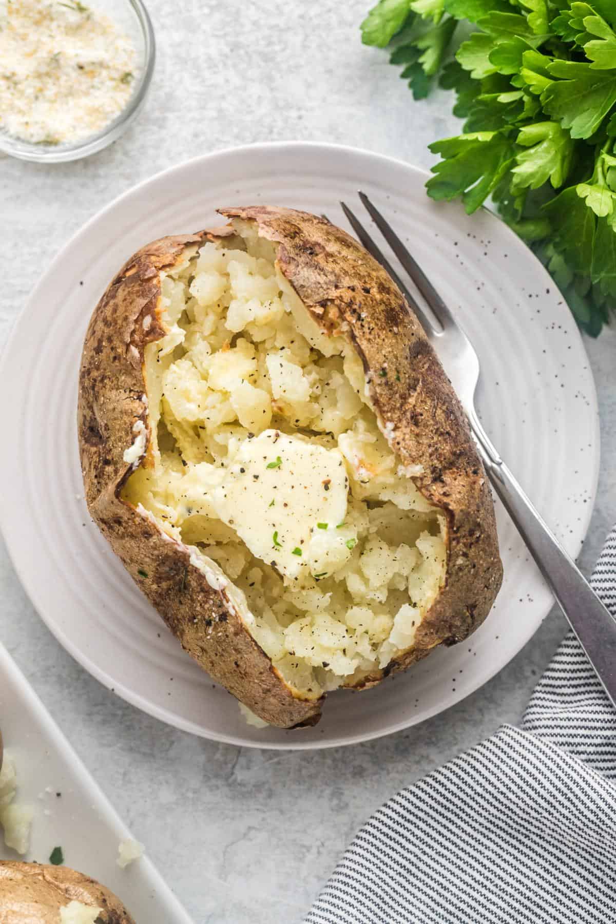 Overhead view of a fluffy baked potato with a pat of melting butter, cracked open and seasoned with black pepper and herbs, served on a white plate with a fork