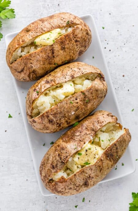 Three baked potatoes on a white tray cut open with butter inside