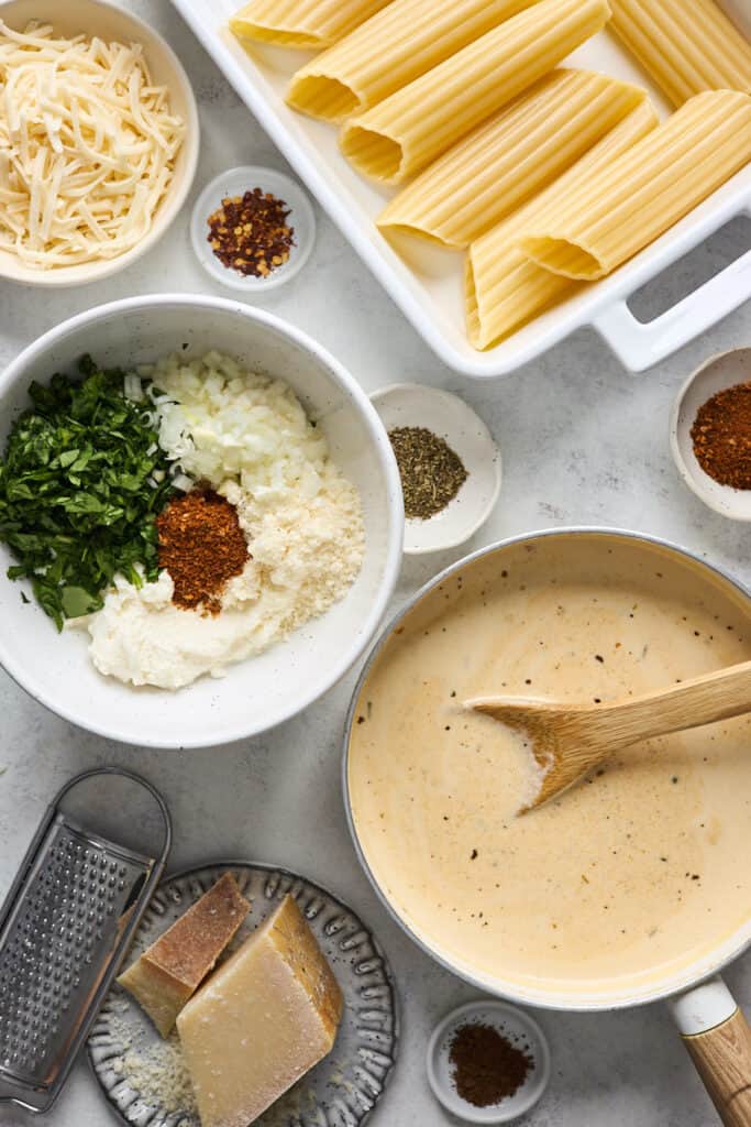 Cheese sauce, spinach stuffing ingredients, manicotti shells and red pepper flakes on a white surface