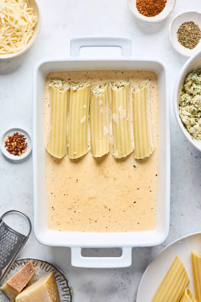 Manicotti being layered in a white baking pan with a cream sauce on the bottom