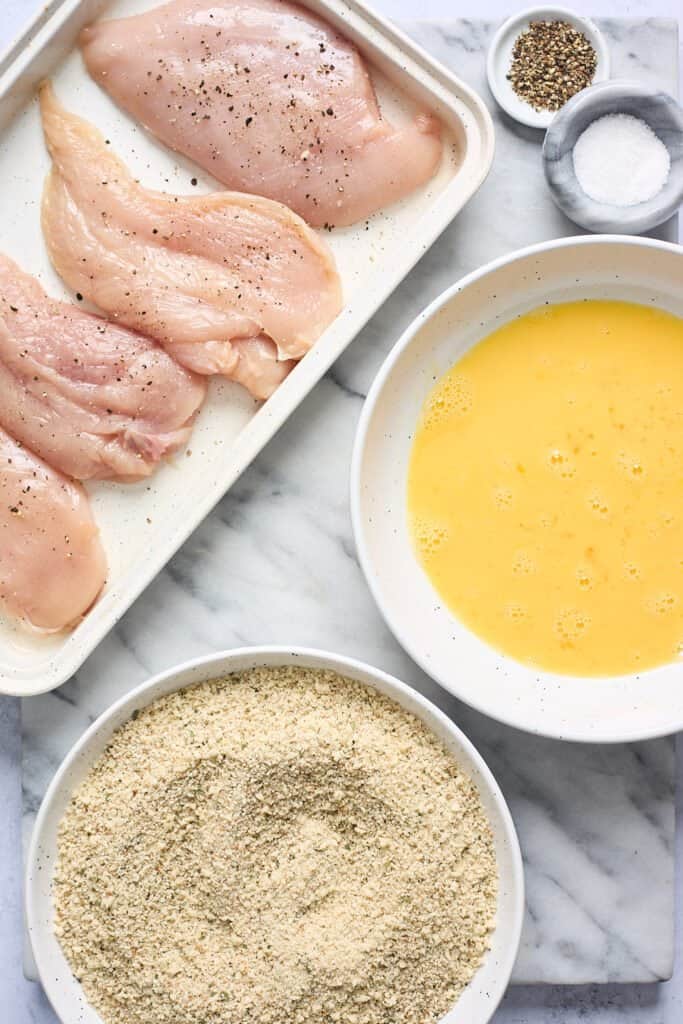 Chicken breasts on a white platter with breadcrumbs and eggs nearby to batter