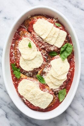 Chicken cutlets in a white pan with marinara sauce on top and mozzarella and basil before baking