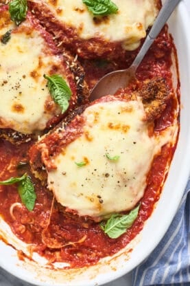 A close up of chicken parmesan being scooped out of white baking pan to serve