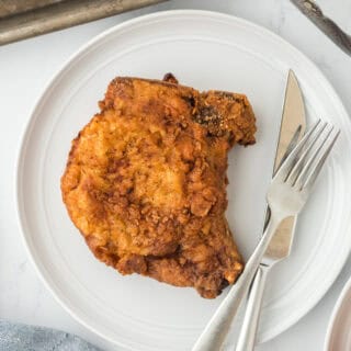 A classic fried pork chop on a white plate with a knife and fork ready to enjoy