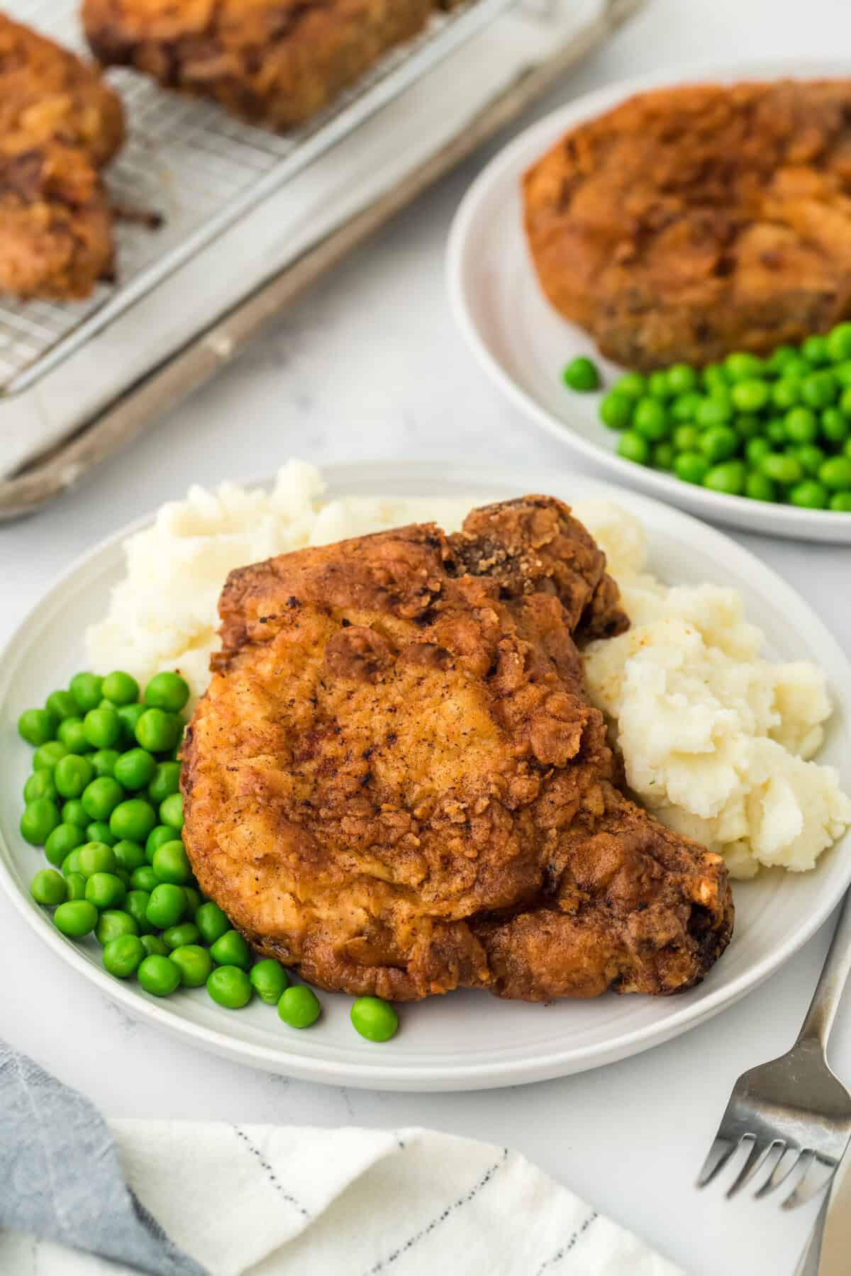 The best fried pork chops recipe on a white plate served with green peas and mashed potatoes with another plate in the white background