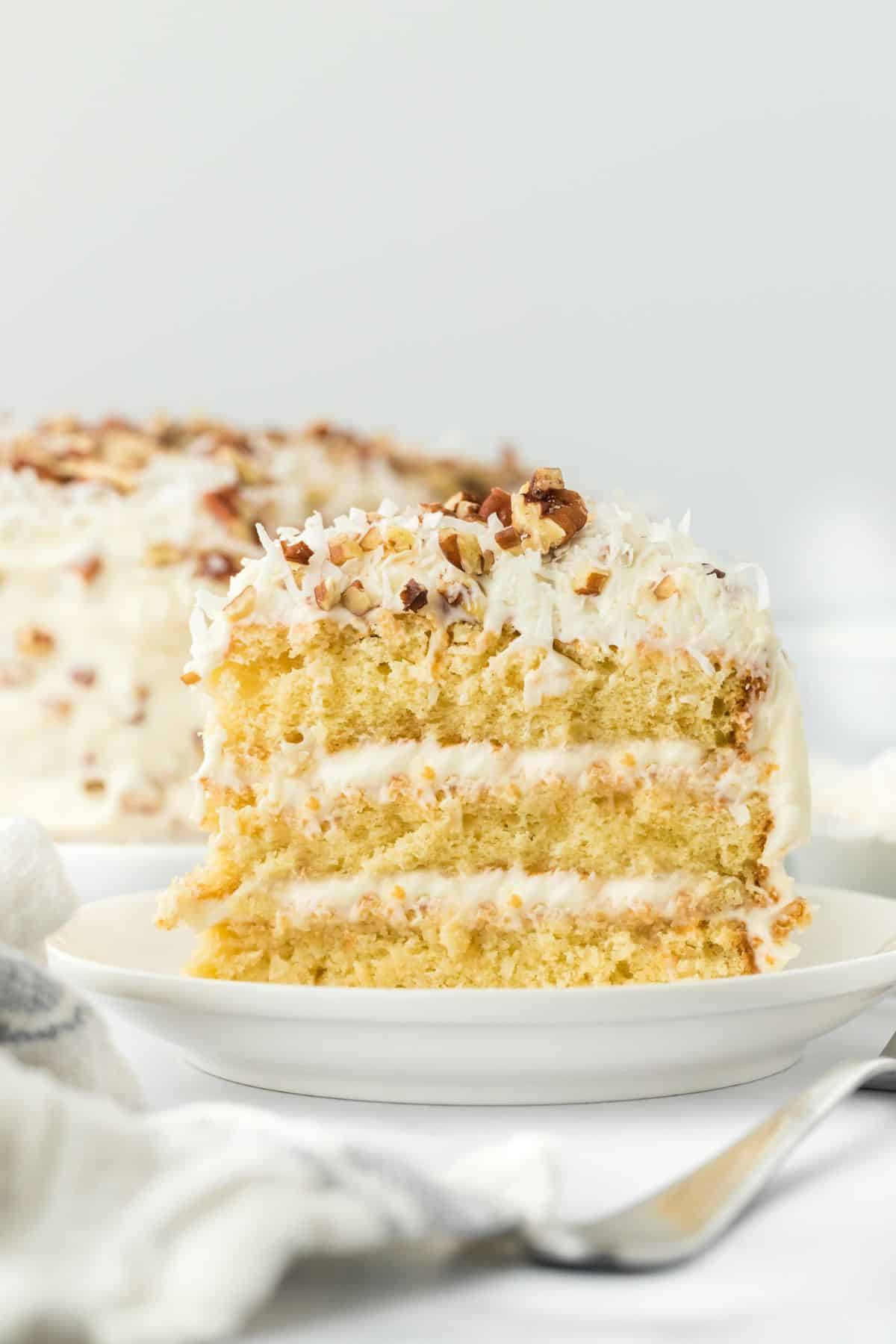 A single slice of Italian cream cake on a plate, showing multiple layers of moist cake and creamy frosting, with the rest of the cake in the background