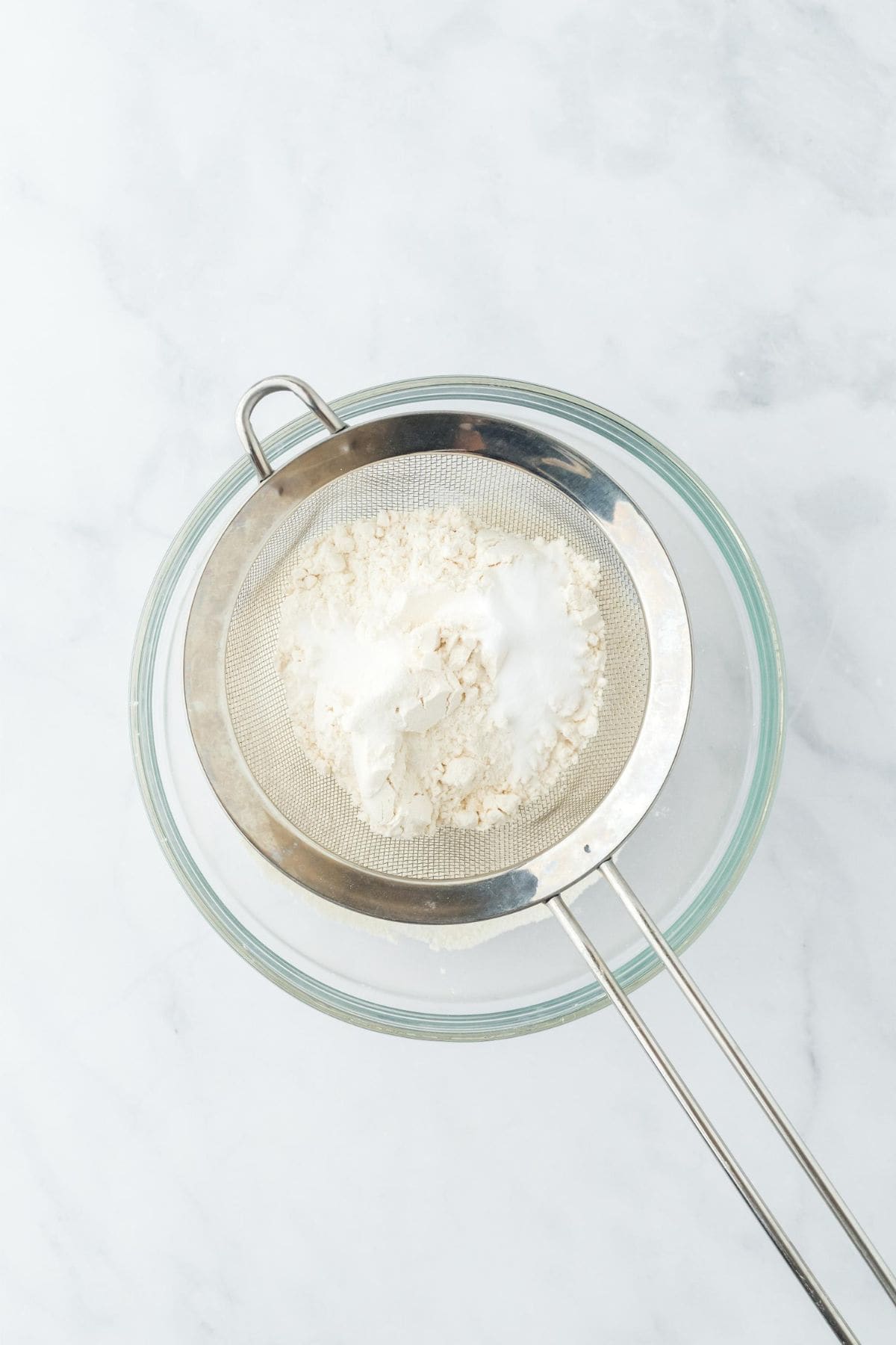 A sifter with dry ingredients being sifted over a glass bowl