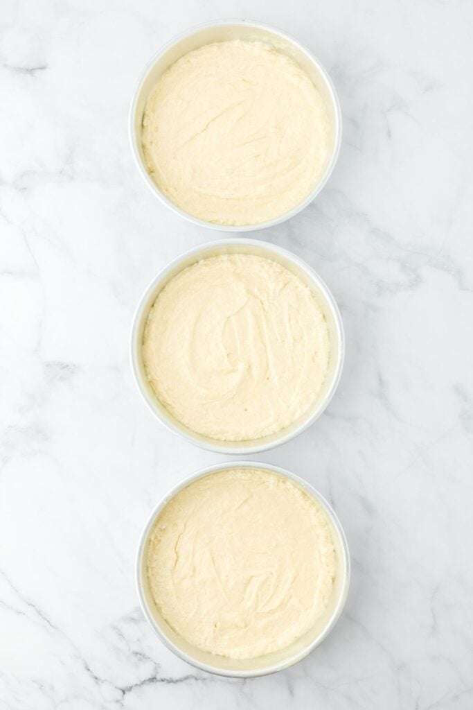 Three round pans filled with cake batter, placed on a marble surface, ready for baking